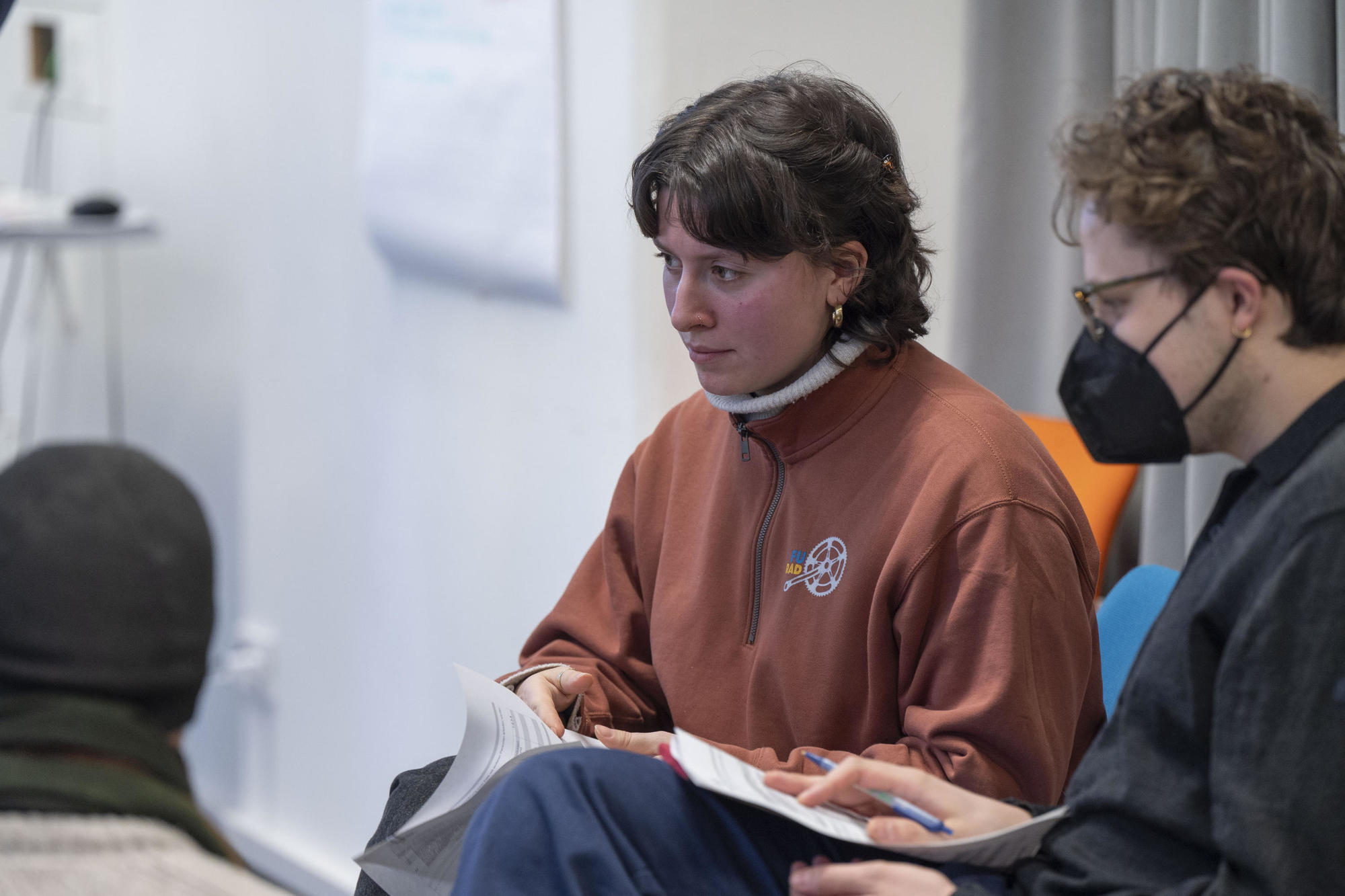Eine Frau in braunem Pullover hält ein Blatt Papier und hört konzentriert zu. Neben ihr sitzt eine Person mit schwarzer Maske und Brille, die Notizen macht. Der Raum wirkt wie ein Seminar- oder Workshopraum.
