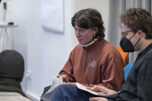 Eine Frau in braunem Pullover hält ein Blatt Papier und hört konzentriert zu. Neben ihr sitzt eine Person mit schwarzer Maske und Brille, die Notizen macht. Der Raum wirkt wie ein Seminar- oder Workshopraum.