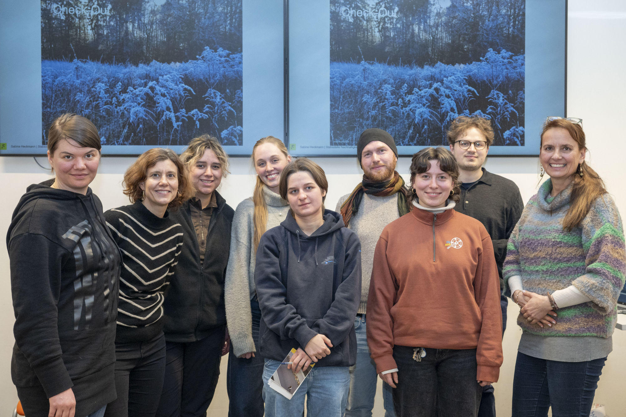Eine Gruppe von neun Personen steht in einem Raum und lächelt in die Kamera. Im Hintergrund sind zwei Bildschirme mit einer winterlichen Landschaft zu sehen. Die Personen tragen verschiedene lässige Outfits, darunter Hoodies, Pullover und Schals.