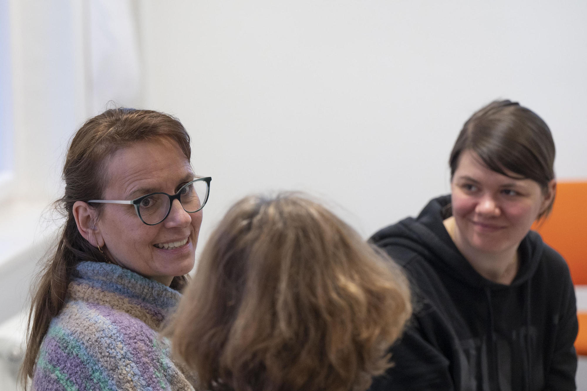 Drei Personen sitzen in einem Raum und unterhalten sich. Eine Frau mit Brille und buntem Strickpullover lächelt in die Kamera. Eine andere Frau in schwarzem Hoodie lächelt ebenfalls. Im Vordergrund ist der Rücken einer Person mit braunen Haaren zu sehen.