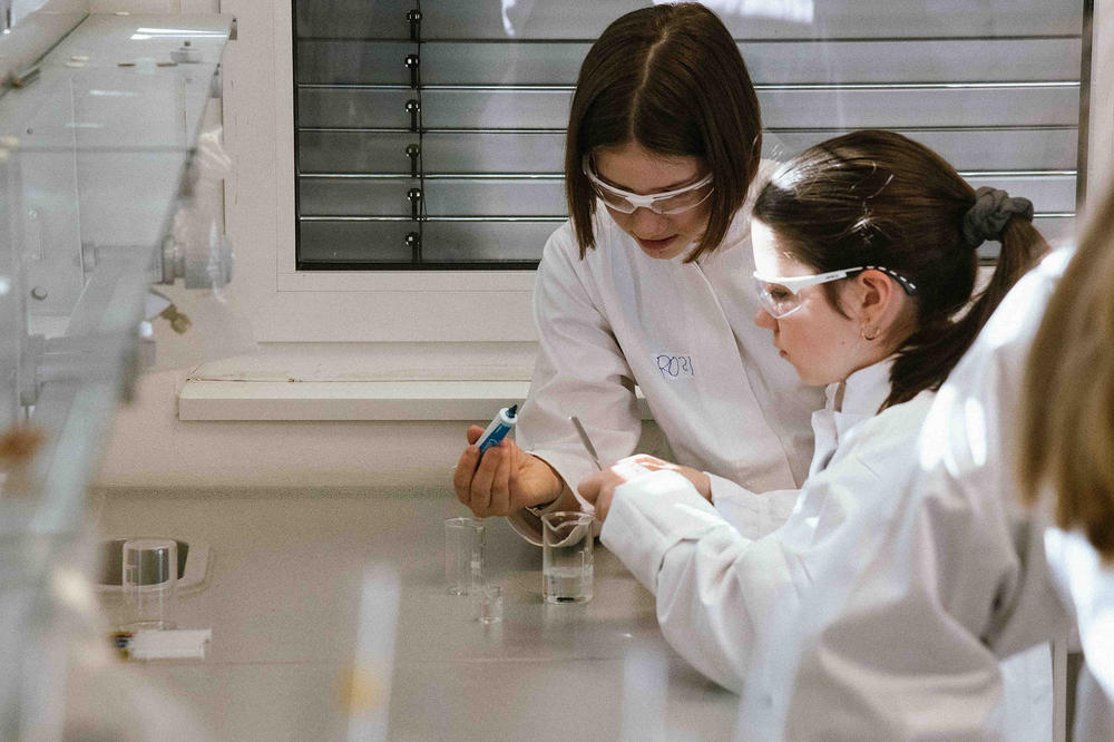 Zwei Mädchen im Laborkittel und mit Schutzbrillen führen gemeinsam ein Experiment durch. Eine hält eine Pipette mit blauer Flüssigkeit, die andere rührt konzentriert in einem Becherglas. Sie arbeiten konzentriert an einem Labortisch.