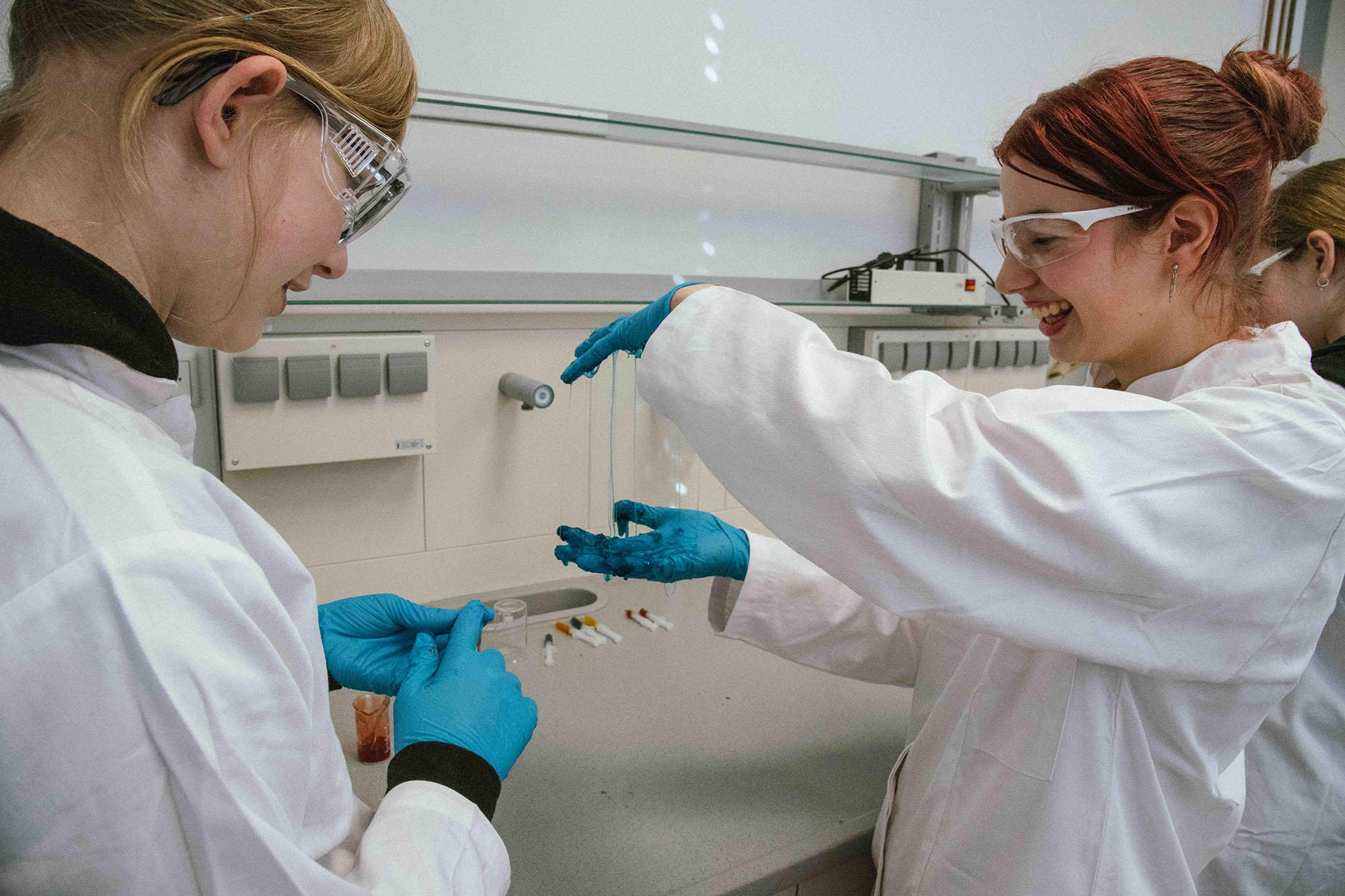 Zwei Mädchen in Laborkitteln und Schutzbrillen experimentieren lachend mit einer schleimartigen Substanz. Beide tragen blaue Schutzhandschuhe, eine hält die Substanz in die Höhe, während die andere interessiert zuschaut.