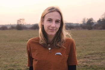 Britt Köhler, eine junge Frau mit schulterlangen blonden Haaren steht leicht lächelnd auf einer offenen Wiese bei sanftem Abendlicht. Sie trägt ein rostbraunes T-Shirt mit einem Dachs-Aufnäher und mehreren Ketten. Im Hintergrund sind Büsche und Bäume zu s