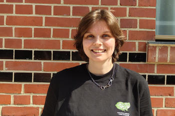 Jule Detmers, eine junge Frau mit kurzen, braunen Haaren, steht lächelnd vor einer Backsteinwand mit schwarzen Ziersteinen. Sie trägt ein schwarzes T-Shirt mit grünem Aufdruck, eine Halskette und hat die Hände in den Hosentaschen. Die Sonne scheint hell.