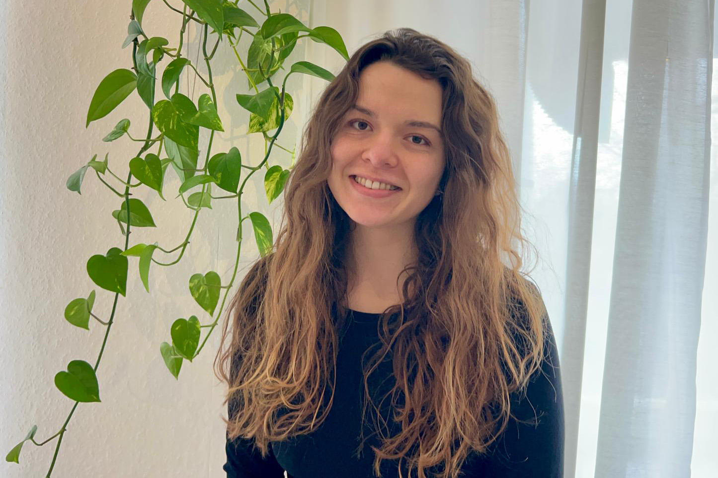 Theresa Langer, eine junge Frau mit langen, welligen braunen Haaren steht lächelnd vor einem hellen Vorhang. Neben ihr hängt eine grüne Rankpflanze in einem weißen Topf an der Wand. Das Licht wirkt weich und natürlich.