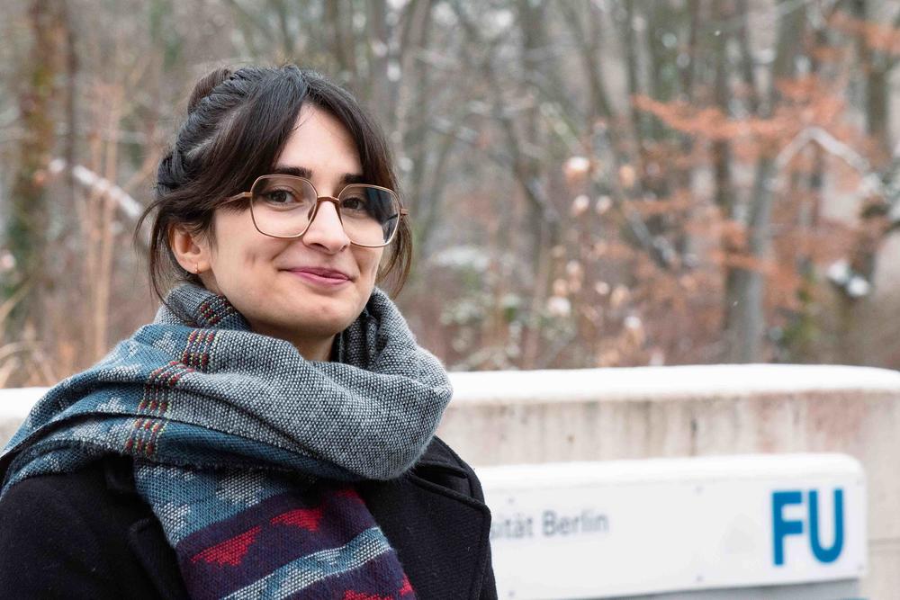 Maryam Keshavarz, dunkle Haaren und Brille, steht im Freien vor einem Schild der Freien Universität Berlin. Sie trägt einen Schal und einen dunklen Mantel. Im Hintergrund sind Bäume mit leichtem Schneefall zu sehen. Sie lächelt und blickt in die Kamera.