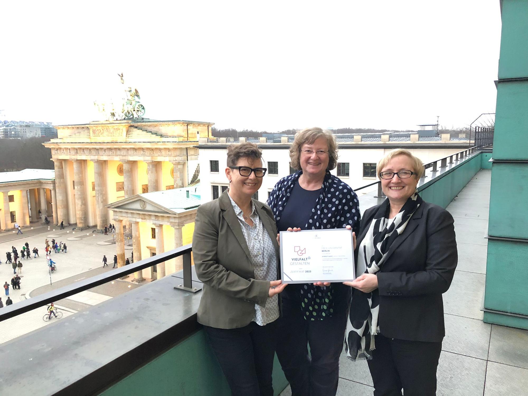 Audit Zertifikatsverleihung: Gabriele Rosenstreich, Dr. Ute Zimmermann und Prof. Dr. Verena Blechinger stehen nebeneinander mit dem Brandenburger Tor im Hintergrund. Sie halten gemeinsam das Audit Zertifikat und lächeln.